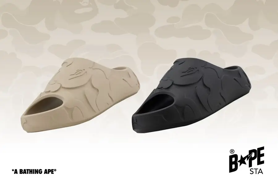 Image showing two BAPE STA slip-on slides in sand beige and matte black, featuring embossed camouflage graphics and signature ape-head detailing against a tonal camo backdrop