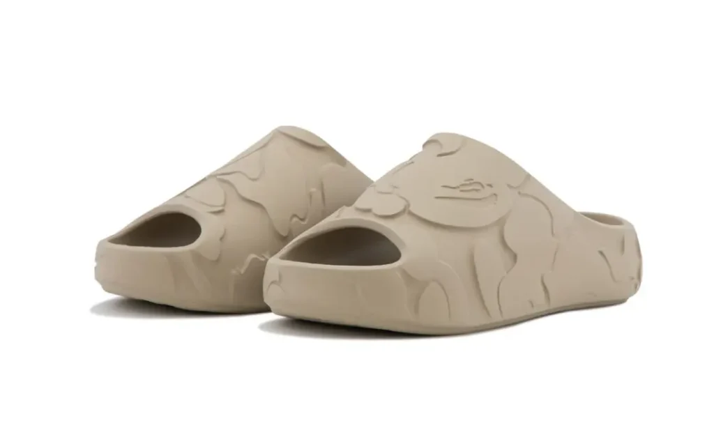 Pair of sand beige BAPE STA foam slides shown in angled studio view with textured camouflage detailing and chunky slip-on proportion