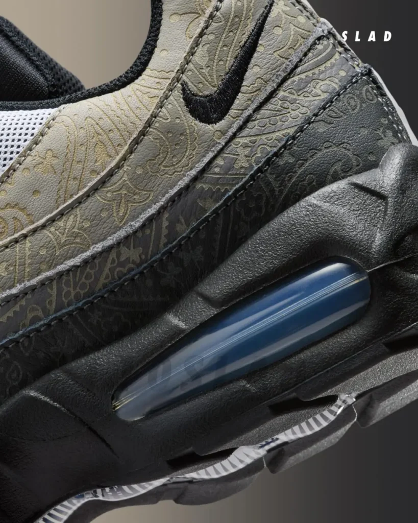 Macro detail shot of the Nike Air Max 95 “Light Charcoal,” focusing on the layered sidewall construction and visible Air cushioning unit. The close-up reveals tonal light charcoal and dark grey overlays finished with an intricate paisley-style etched pattern across the signature Air Max 95 waves, contrasted against black embroidered Swoosh detailing and textured black midsole tooling. The exposed Air unit introduces a subtle translucent blue tint, while the combination of decorative surface treatment and sculpted performance elements gives the sneaker a more elevated and premium interpretation of the classic running sil