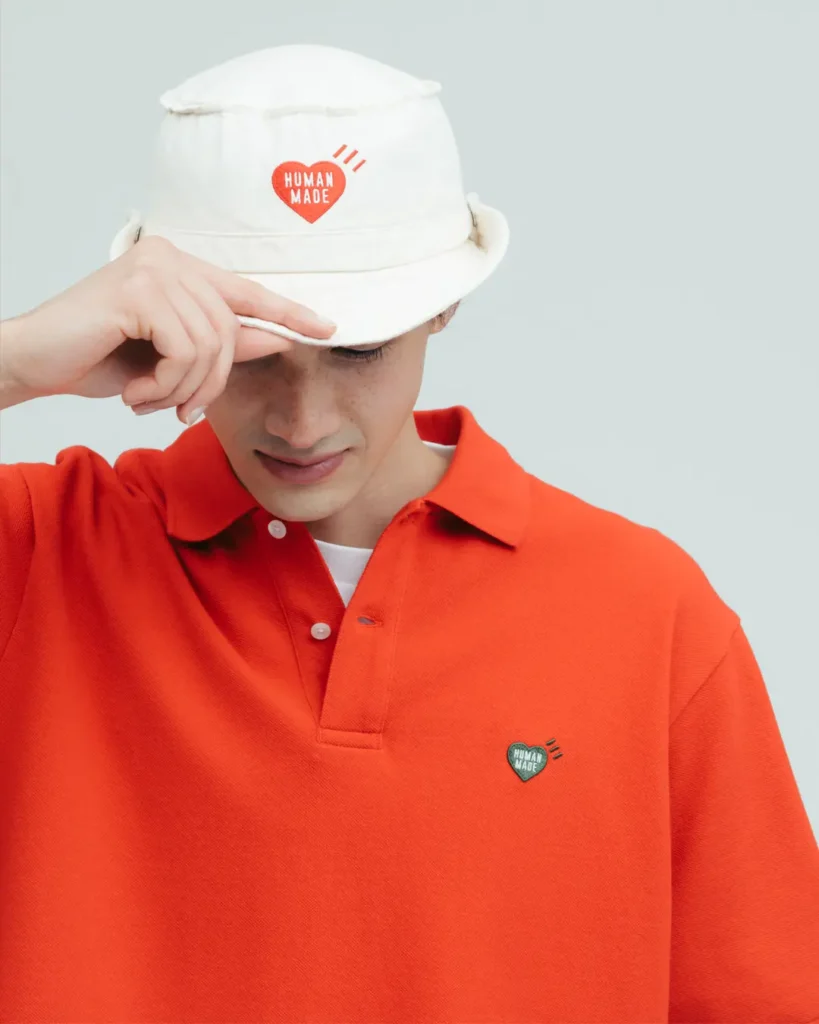 Close-up portrait of a model adjusting a cream Human Made bucket hat while wearing a bright red oversized polo shirt with embroidered heart logo detail