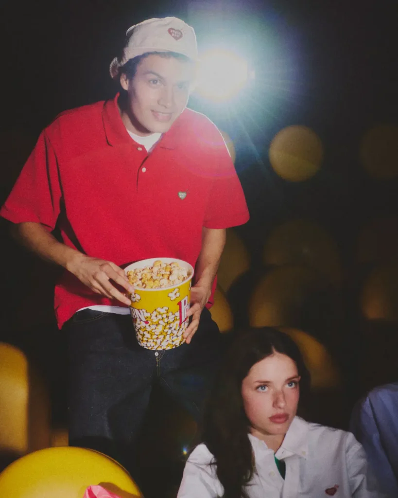 Model in a bright red Human Made polo shirt and white bucket hat holding a popcorn bucket under dramatic cinema lighting during a retro-inspired fashion campaign