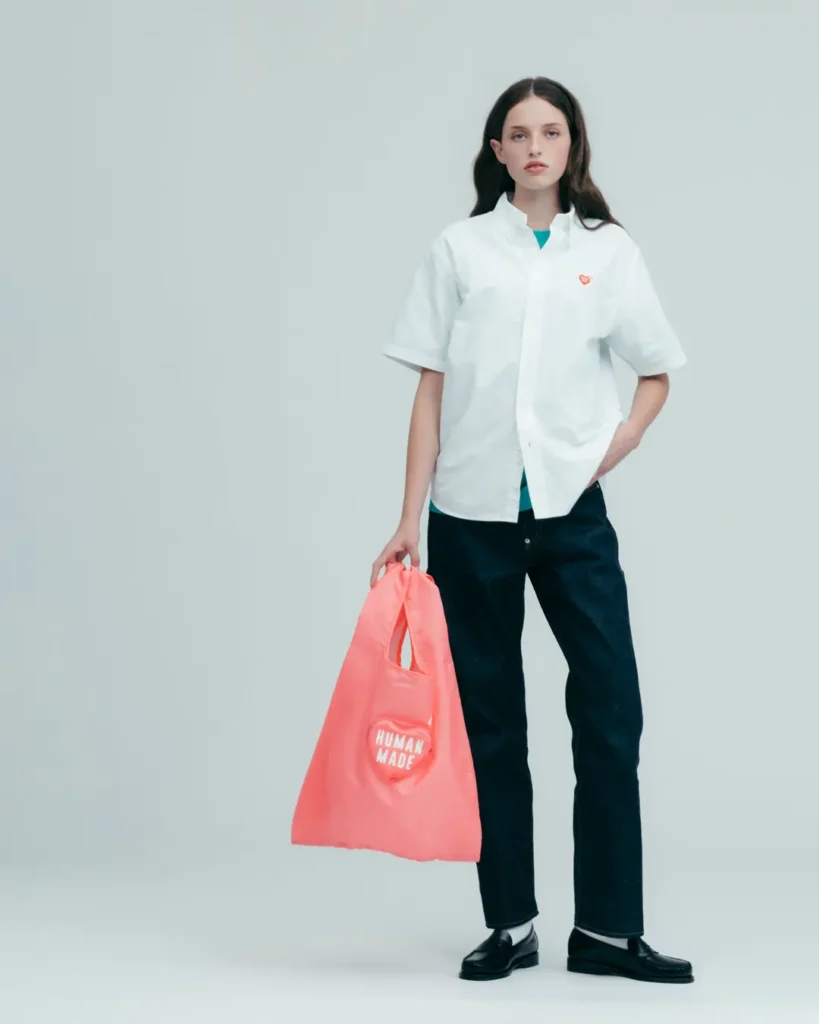 Female model wearing a white Human Made short-sleeve shirt, dark denim, and loafers while holding a pink logo tote bag against a minimalist light gray studio backdrop