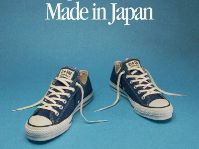 A clean campaign-style image featuring the Gramicci x Converse “Original All Star Made in Japan” sneakers against a vivid sky-blue backdrop. The low-top canvas shoes appear in deep indigo blue with white laces, rubber toe caps, and vintage-style detailing, positioned casually with untied laces spread outward. Large retro serif typography reading “Original All Star Made in Japan” dominates the upper portion of the composition, while Gramicci and Converse branding sits subtly in the lower right corner, emphasizing the collaboration’s heritage-focused Japanese craftsmanship aesthetic
