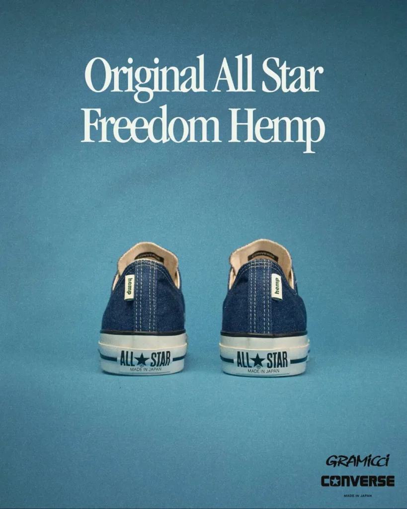 Rear-view campaign image of the Gramicci x Converse “Original All Star Freedom Hemp” shoe set against a bright sky-blue studio backdrop. The low-top shoes feature textured indigo hemp uppers with contrast white stitching, classic white rubber midsoles, and bold “ALL STAR Made in Japan” heel branding. Small hemp tags appear along the heel seams, reinforcing the eco-conscious material focus, while oversized serif typography reading “Original All Star Freedom Hemp” floats above the pair. Gramicci and Converse logos are positioned subtly in the lower corner, giving the view a vintage Japanese catalog-inspired feel