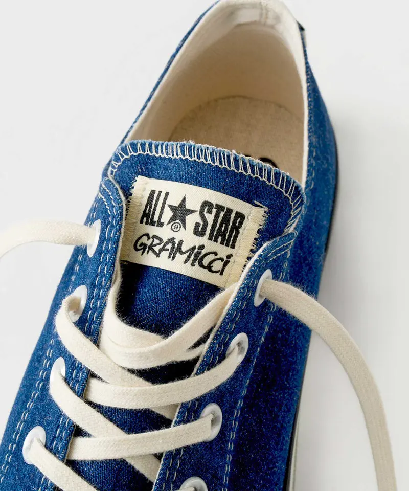 Close-up detail image of the Gramicci x Converse shoe tongue and upper construction in textured indigo hemp fabric. The shot highlights the co-branded “ALL★STAR Gramicci” woven tongue label, thick off-white laces, exposed stitching around the collar, and contrast seam detailing throughout the upper. Captured against a clean light gray backdrop, the composition emphasizes the collaboration’s handcrafted Made in Japan aesthetic and rugged outdoor-inspired material treatment