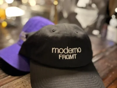 Black Moderno x FRGMT baseball cap photographed on a rustic wooden table alongside a purple colorway, featuring minimalist white embroidery and Hiroshi Fujiwara’s signature understated streetwear aesthetic