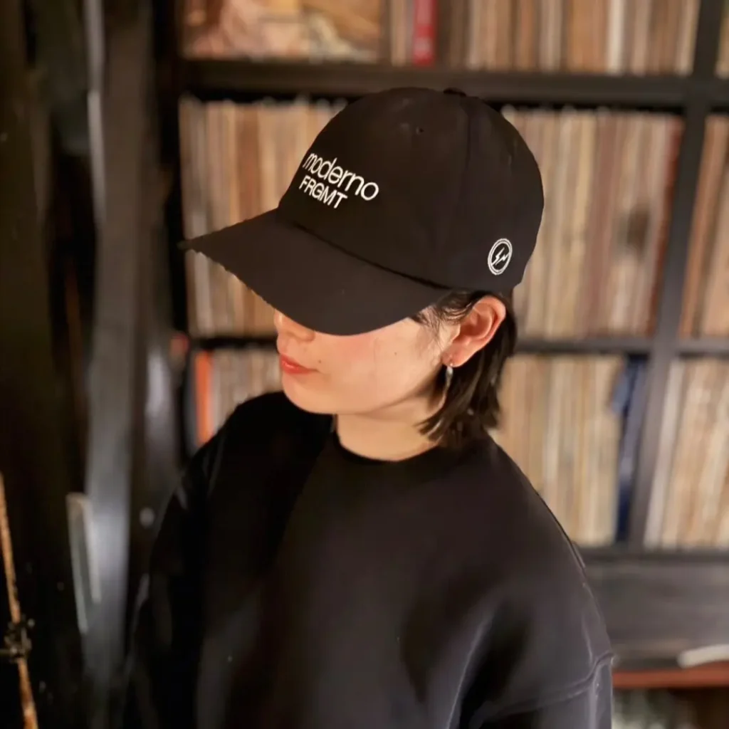 Black Moderno x FRGMT baseball cap photographed on a rustic wooden table alongside a purple colorway, featuring minimalist white embroidery and Hiroshi Fujiwara’s signature understated streetwear aesthetic