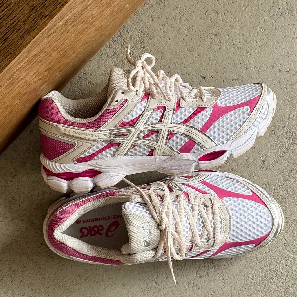 Overhead lifestyle shot of the ASICS GEL-CUMULUS 16 featuring a white engineered mesh upper with cream overlays and vivid pink accents, placed on a textured floor beside a wooden bench