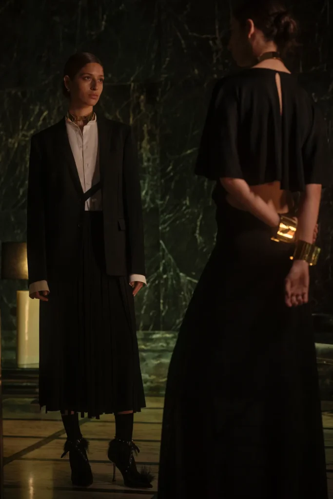 Minimalist black eveningwear featuring a pleated skirt, sharp blazer tailoring, gold jewelry accents, and soft cinematic lighting within a marble-clad interior