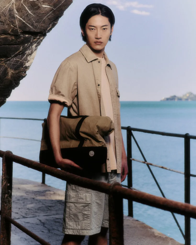 A model stands along a weathered coastal walkway overlooking calm blue water, framed by rugged rock formations and rusted industrial railings that evoke a quiet maritime atmosphere. He wears a relaxed sand-toned short-sleeve overshirt layered over a pale neutral tee, paired with textured utility shorts in a muted stone shade. Carried under the arm is a structured technical duffel bag featuring dark contrast panels and subtle Stone Island branding. The composition merges utilitarian resortwear with refined coastal minimalism, emphasizing soft natural palettes, functional layering, and the brand’s understated approach to luxury travel and outdoor performance