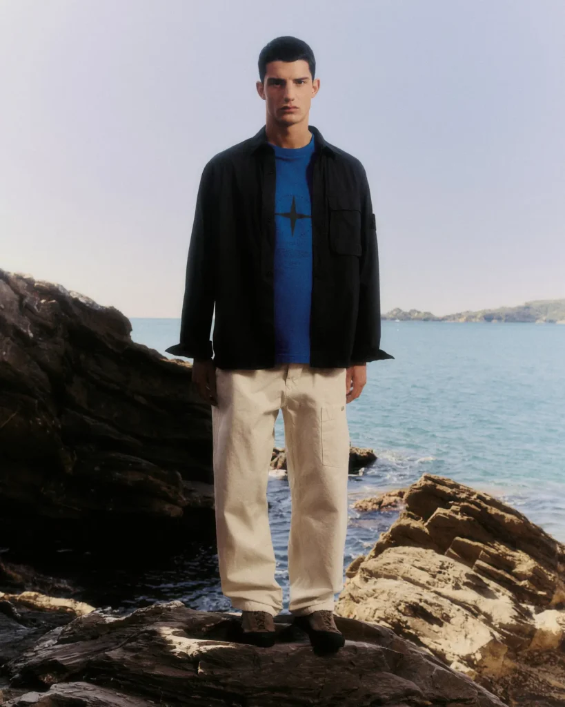 A model stands atop rugged coastal rocks overlooking calm turquoise water beneath a pale open sky, dressed in relaxed Stone Island essentials that balance utility with understated elegance. The look combines a dark overshirt layered over a vivid cobalt blue graphic tee featuring the brand’s compass-inspired motif, paired with loose off-white utility trousers and dark footwear suited for coastal terrain. The composition contrasts sharp monochromatic layering against the bright maritime backdrop, reinforcing Stone Island’s fusion of technical menswear, minimal resort styling, and refined outdoor functionality