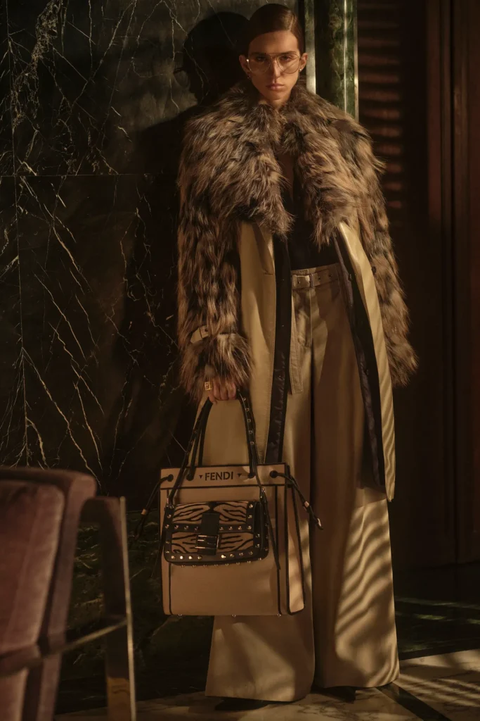 Model wearing a fur-trimmed neutral trench coat with wide-leg trousers and a zebra-accented Fendi tote bag, blending Roman luxury with modern safari-inspired textures