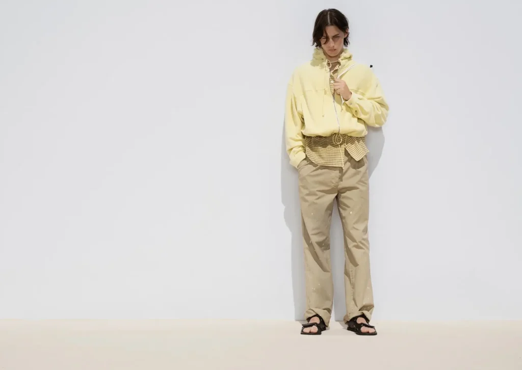 Model wearing a pale yellow oversized zip jacket layered over a gingham shirt with relaxed beige trousers and black sandals against a soft neutral backdrop in AURALEE Spring/Summer 2026 campaign imagery