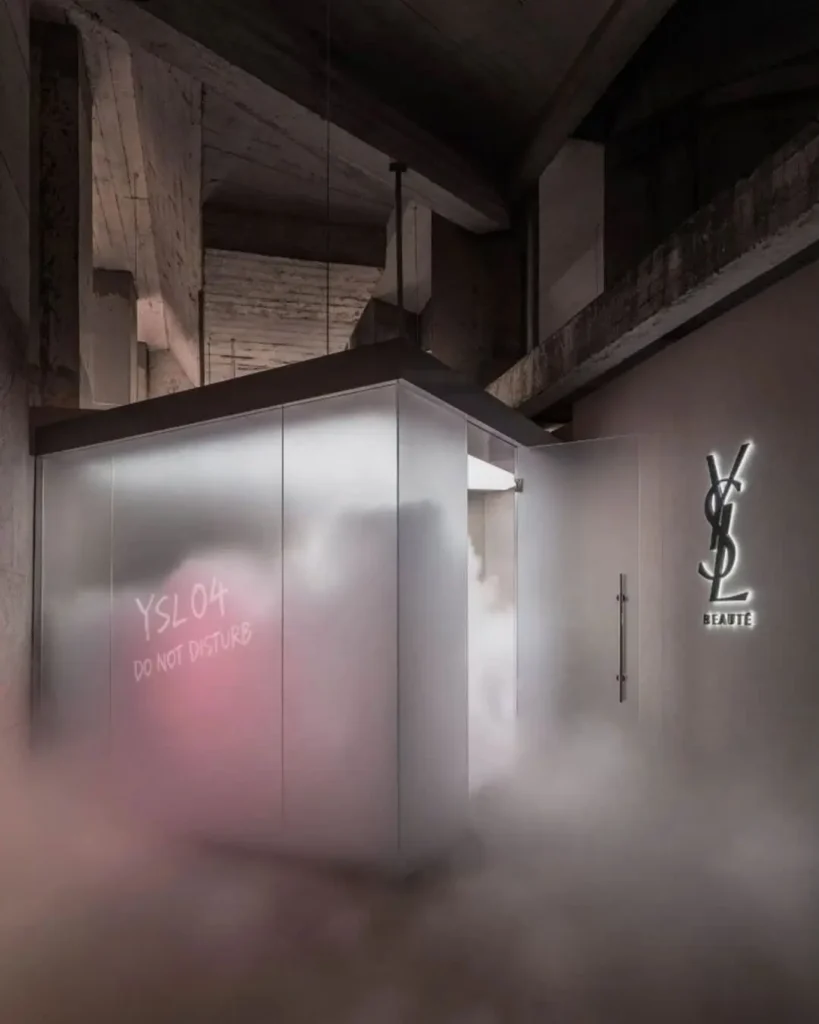 Immersive room installation inside the YSL Beauty LoveNude Hotel pop-up in Shanghai, featuring frosted translucent walls marked with “YSL 04 Do Not Disturb,” illuminated YSL Beauté signage, theatrical low-lying fog, and exposed industrial concrete architecture that blends cinematic hospitality themes with avant-garde luxury beauty retail design