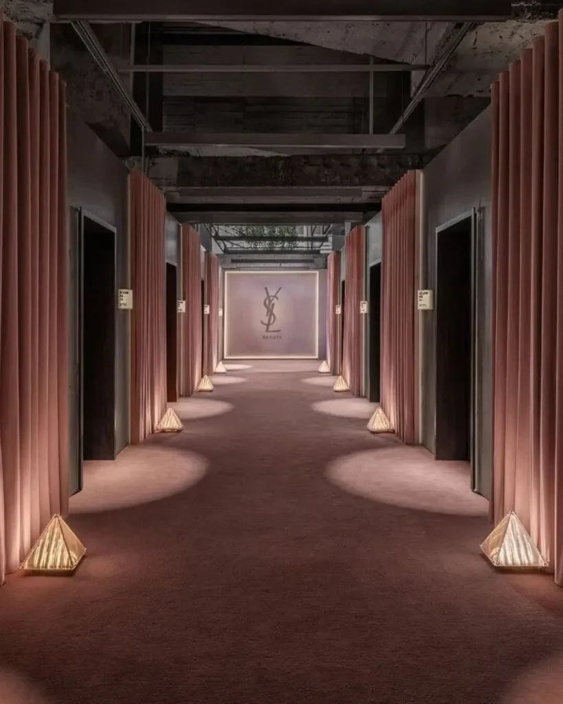 Hotel-inspired corridor inside the YSL Beauty LoveNude Hotel activation in Shanghai, featuring symmetrical rows of blush pink drapery-lined doorways, softly illuminated carpeting, sculptural floor lighting, exposed industrial ceilings, and a glowing YSL Beauté monogram at the far end that reinforces the immersive hospitality-meets-luxury-retail atmosphere