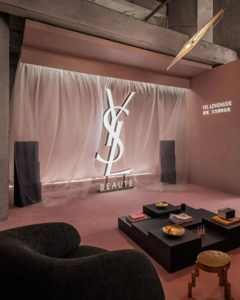 Lounge-style interior inside the YSL Beauty LoveNude Hotel pop-up in Shanghai, featuring illuminated YSL Beauté signage against sheer blush drapery, plush pink carpeting, sculptural black seating, low minimalist tables with beauty products and books, and raw concrete architecture that blends intimate hospitality atmospheres with immersive luxury retail design