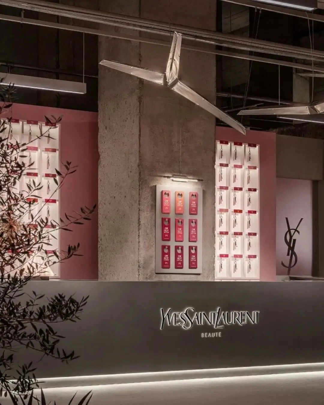 Reception-style interior inside the YSL Beauty LoveNude Hotel activation in Shanghai, featuring illuminated Yves Saint Laurent Beauté branding, raw concrete architecture, blush-toned product walls displaying room keys and beauty items, sculptural metallic lighting fixtures, and immersive hospitality-inspired retail design blending industrial minimalism with luxury beauty aesthetics
