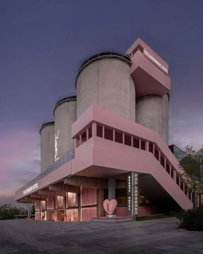 Exterior view of the YSL Beauty LoveNude Hotel activation in Shanghai, showcasing towering raw concrete silos wrapped with blush pink architectural interventions, elevated stairways, glowing YSL branding, and hospitality-inspired installations that merge industrial architecture with immersive luxury beauty retail design
