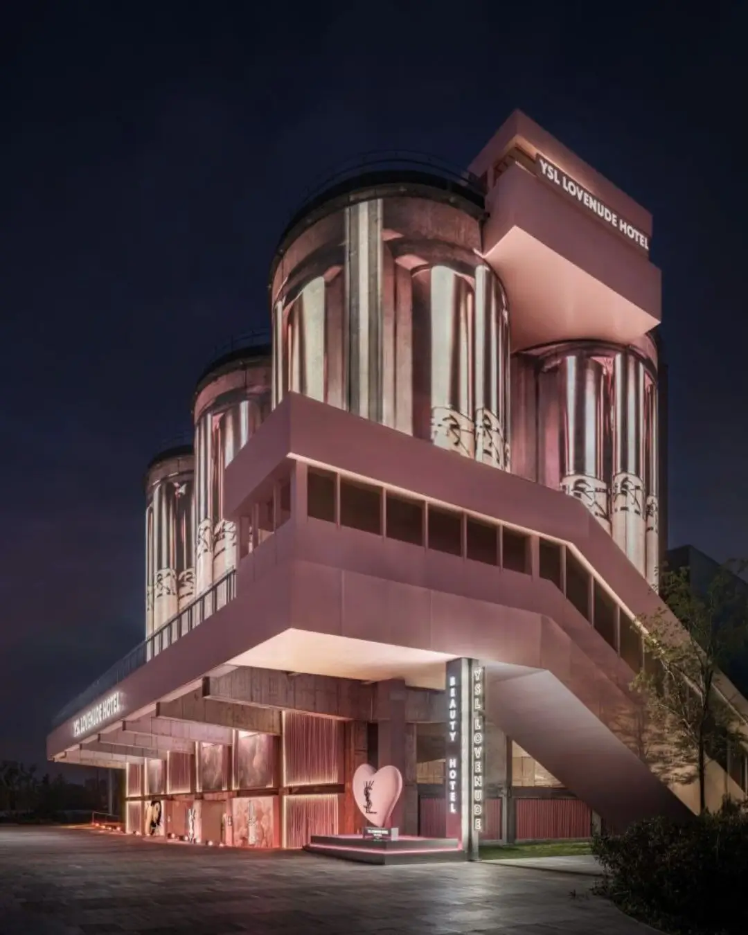 Nighttime exterior view of the YSL Beauty LoveNude Hotel pop-up in Shanghai, featuring illuminated cylindrical industrial silos wrapped in soft pink lighting, elevated modernist walkways, glowing YSL signage, and hospitality-inspired architectural installations that transform the former industrial site into an immersive luxury beauty destination