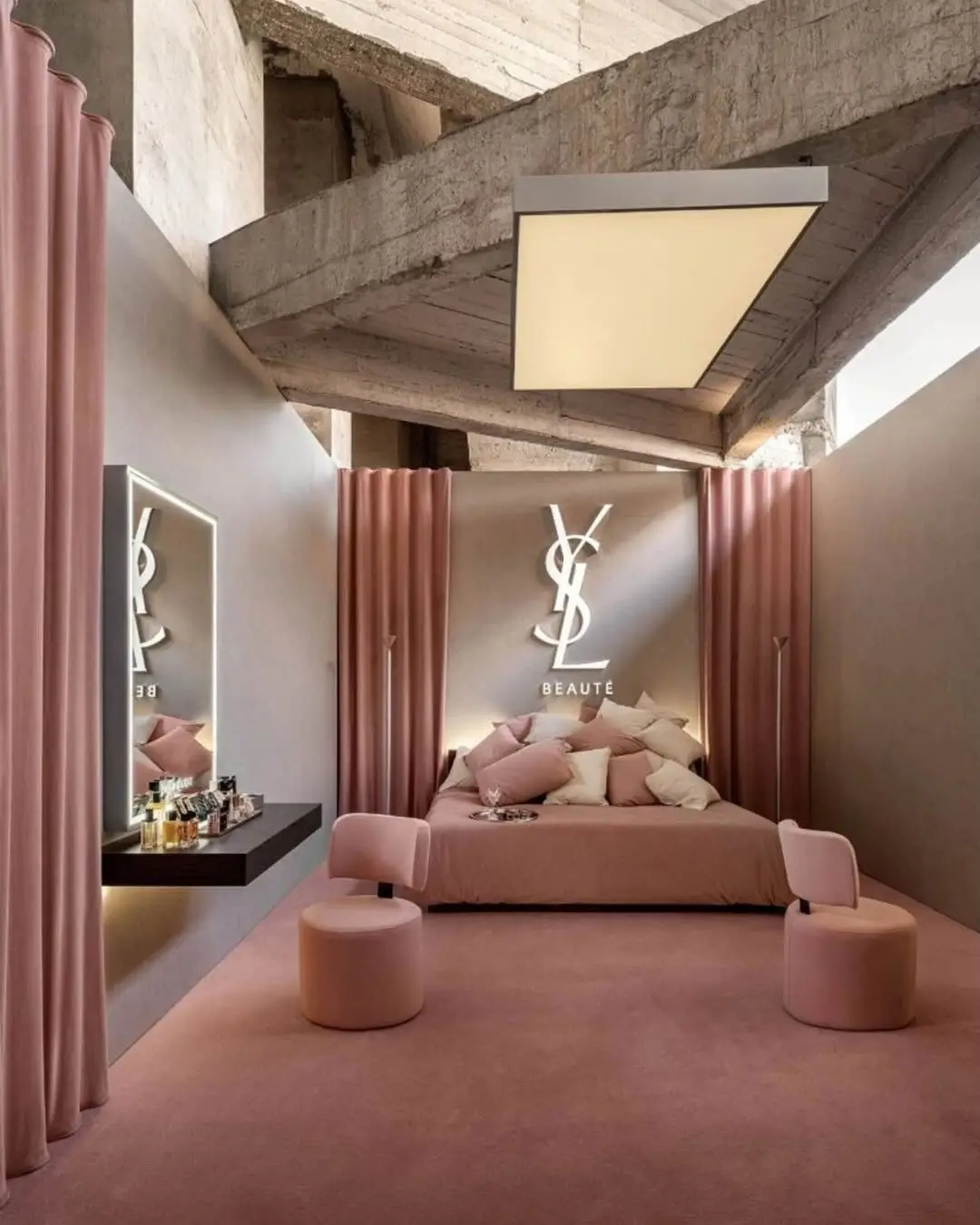 Bedroom-inspired suite inside the YSL Beauty LoveNude Hotel pop-up in Shanghai, featuring blush pink carpeting, sculptural seating, soft draped curtains, fragrance and beauty displays, and an illuminated YSL Beauté monogram set against raw industrial concrete architecture that merges intimate hospitality aesthetics with immersive luxury retail design