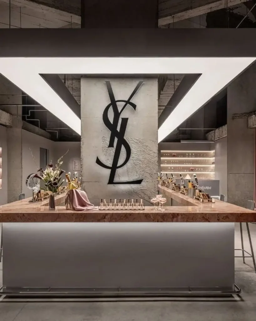 Luxury beauty bar inside the YSL Beauty LoveNude Hotel pop-up in Shanghai, featuring illuminated marble counters lined with mirrors and cosmetics, towering raw concrete pillars marked by a large YSL monogram, blush-toned drapery, and industrial-modern interiors that blend hospitality aesthetics with immersive experiential retail design
