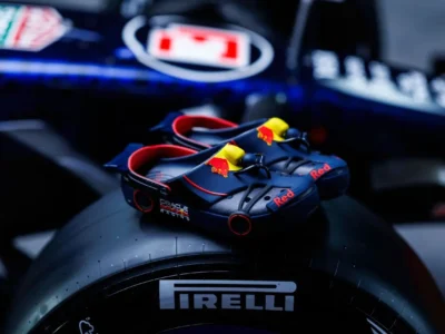 A pair of Oracle Red Bull Racing-inspired clogs rest atop a Pirelli Formula 1 tire, finished in matte navy with vivid red piping, Red Bull iconography, and miniature aerodynamic detailing that echoes the engineering language of an F1 car. The cinematic lighting and blurred paddock backdrop amplify the motorsport aesthetic, making the footwear read less like casual slip-ons and more like collectible racing equipment styled for the grid