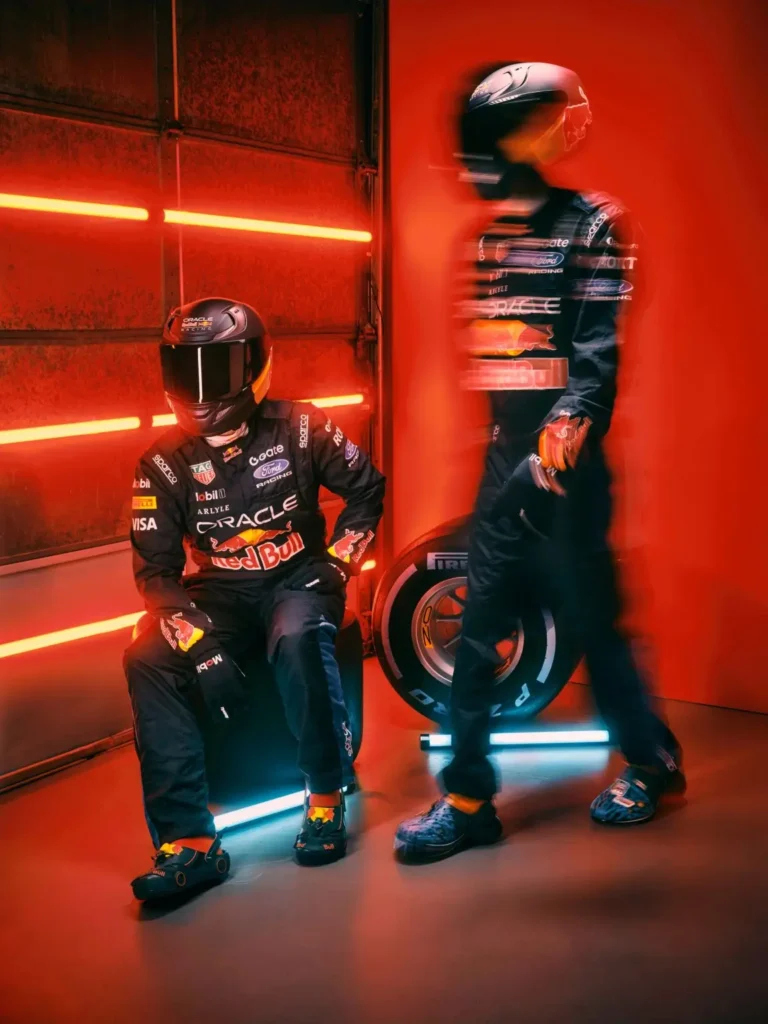 Two helmeted figures in full Oracle Red Bull Racing suits pose inside a neon-lit garage washed in saturated red light, with one seated beside a Pirelli tire while the other moves through the frame in a motion blur. Both wear Formula 1-inspired clogs featuring Red Bull branding, miniature wheel-like detailing, and aerodynamic accents, transforming the footwear into an extension of the racing uniform rather than a casual accessory. The cinematic lighting, industrial backdrop, and blurred movement create the atmosphere of a stylized pit-lane editorial merging motorsport precision with futuristic streetwear energy