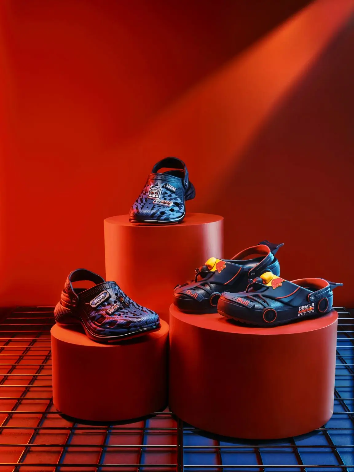 A trio of Oracle Red Bull Racing-inspired clogs and Crocs-style silhouettes are displayed on cylindrical podiums against a saturated red backdrop, styled like a futuristic motorsport showroom installation. The collection mixes aerodynamic detailing, racing graphics, sponsor-style patches, and miniature wheel accents, with navy and electric blue finishes accented by vivid red and yellow touches drawn from Formula 1 pit-lane aesthetics. Dramatic spotlighting and the industrial grid floor elevate the footwear into sculptural design objects, blurring the line between performance gear, collectible merchandise, and conceptual streetwear