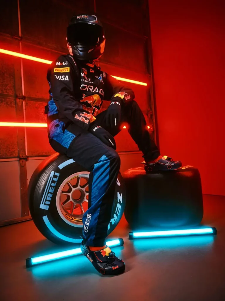 Two helmeted figures in full Oracle Red Bull Racing suits pose inside a neon-lit garage washed in saturated red light, with one seated beside a Pirelli tire while the other moves through the frame in a motion blur. Both wear Formula 1-inspired clogs featuring Red Bull branding, miniature wheel-like detailing, and aerodynamic accents, transforming the footwear into an extension of the racing uniform rather than a casual accessory. The cinematic lighting, industrial backdrop, and blurred movement create the atmosphere of a stylized pit-lane editorial merging motorsport precision with futuristic streetwear energy