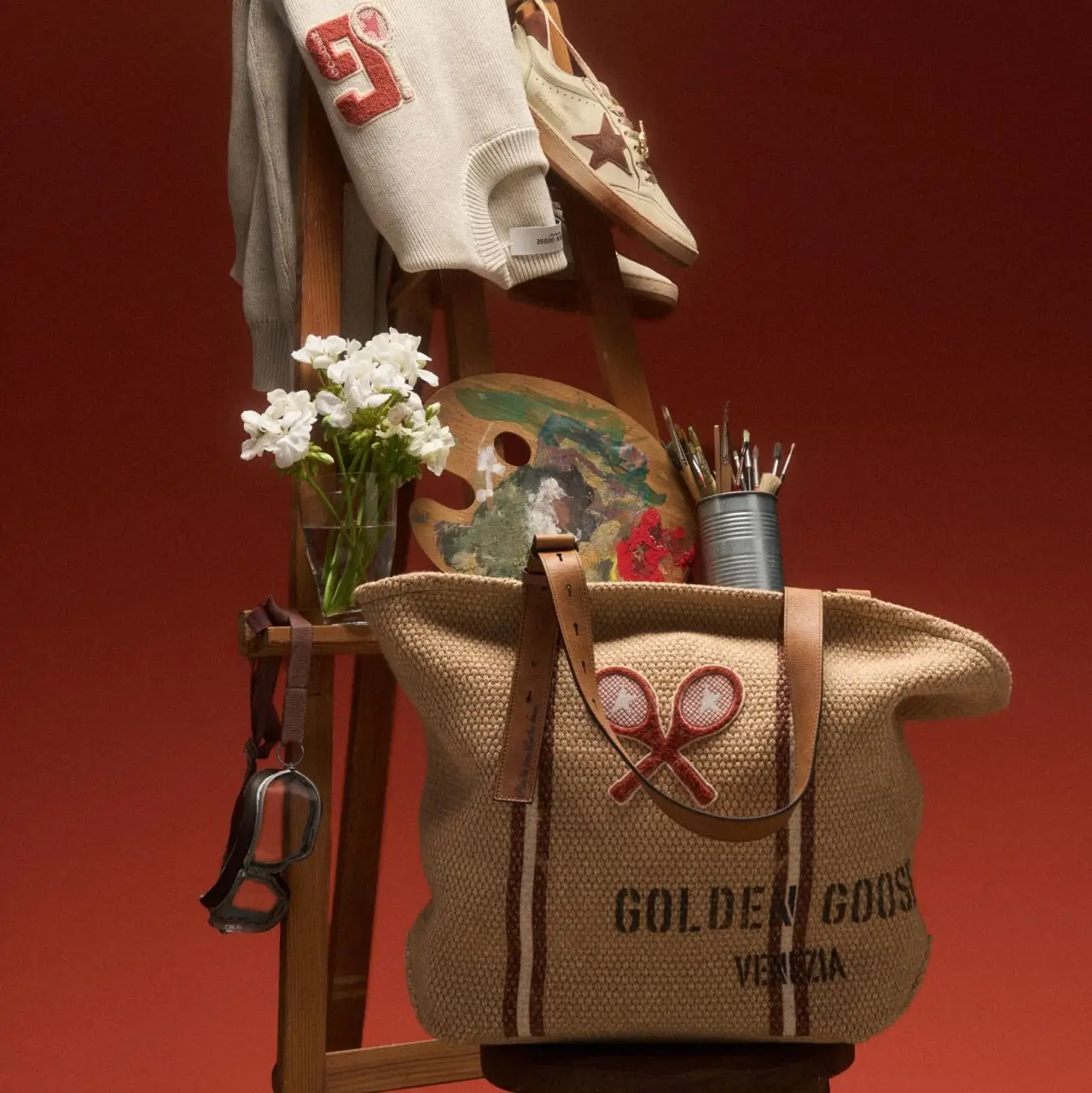 Editorial still life featuring Golden Goose’s sport-inspired collection arranged in an artist’s studio setting. A woven raffia tote with leather straps and a tennis-racket motif sits in the foreground against a deep terracotta backdrop, surrounded by a painter’s palette, brushes, white flowers and vintage-style goggles. Above, a cream knit sweater with varsity-style detailing and distressed Golden Goose sneakers hang from a wooden ladder, blending athletic heritage with artistic expression and handcrafted Italian lifestyle aesthetics