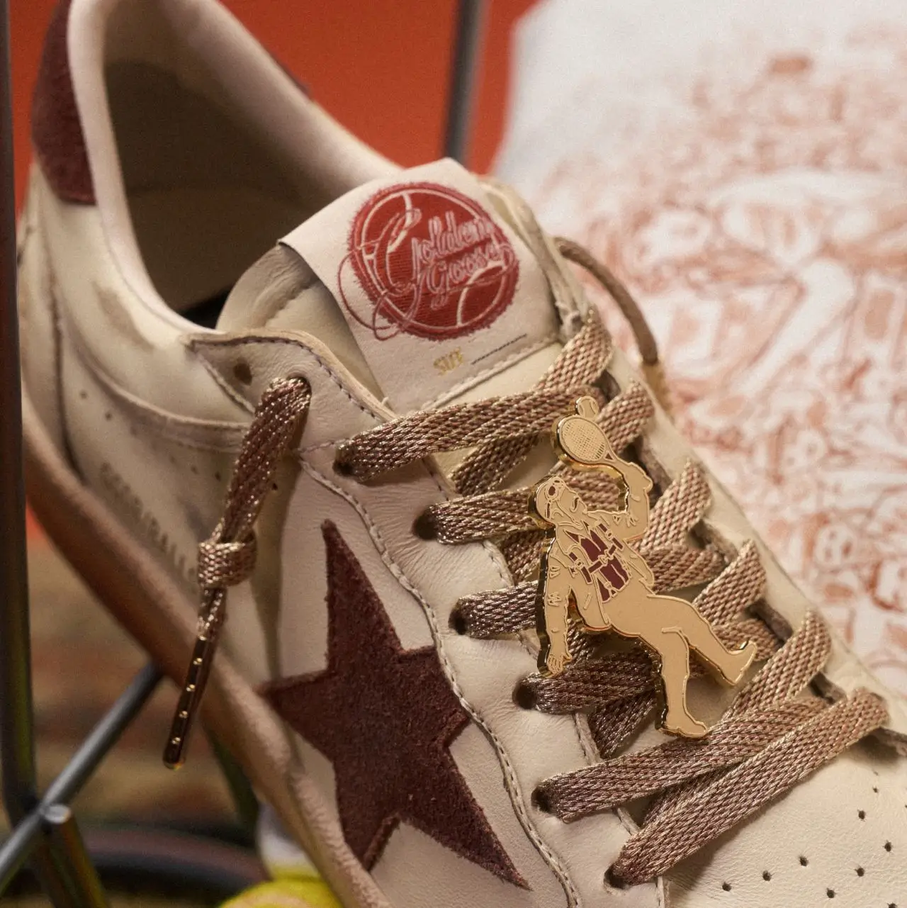 Close-up editorial detail of a Golden Goose sneaker in off-white distressed leather with tonal brown laces and the brand’s signature textured star appliqué at the side. Attached to the laces is a gold-tone charm depicting a vintage tennis player mid-swing, reinforcing the collection’s sport-inspired narrative. Warm terracotta tones and illustrated background elements create an artisanal, nostalgic atmosphere that blends athletic heritage with handcrafted luxury