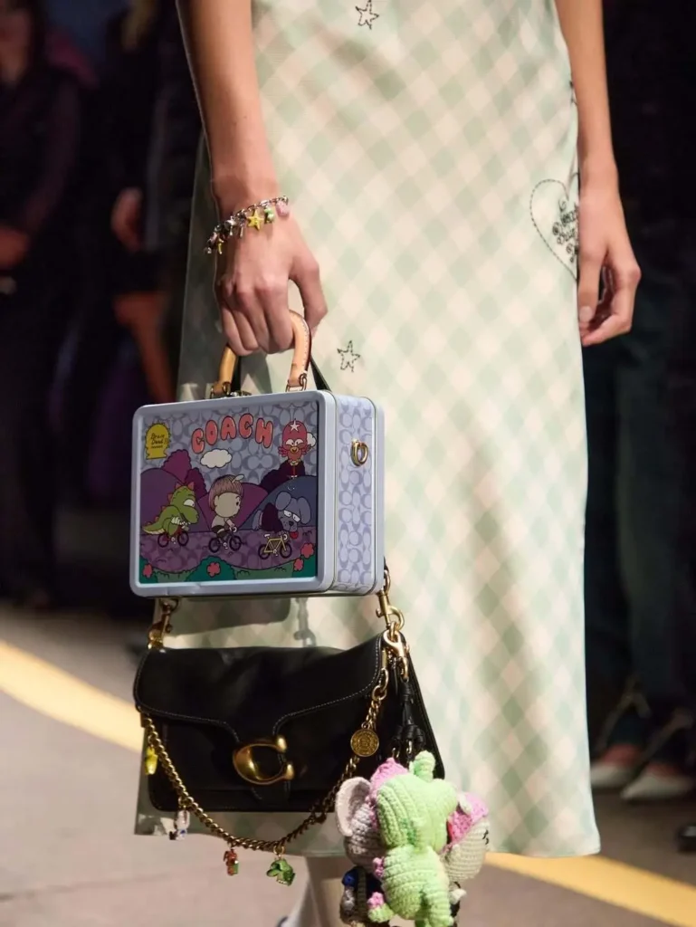 Runway close-up of layered Coach accessories styled with play collectible details. A model in a pale mint gingham slip dress carries a structured graphic case bag featuring illustrated cartoon characters and bold Coach branding, layered above a black leather shoulder bag with gold hardware and chain accents. Crocheted plush charms, miniature hanging ornaments, and colorful bracelet details reinforce the collection’s nostalgic, souvenir-inspired, and youthfully customized aesthetic