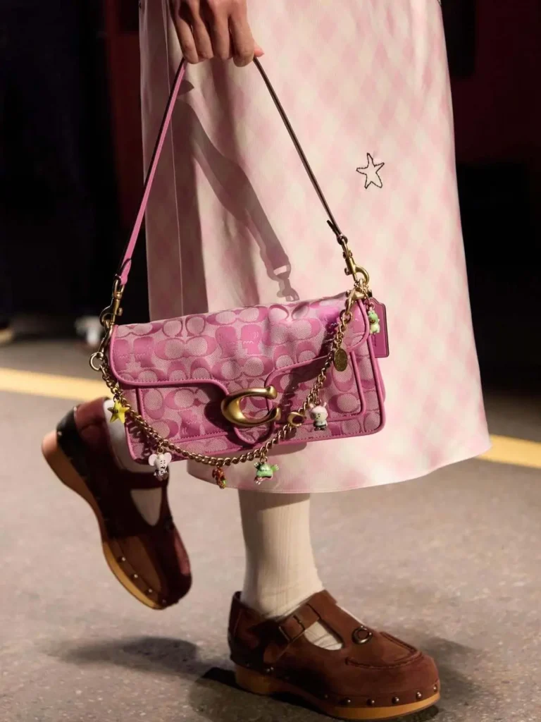 Runway close-up of a pastel pink Coach shoulder bag styled with layered charm customization and playful detailing. The elongated silhouette features tonal monogram jacquard, smooth leather trim, and oversized gold-tone hardware, finished with a draped chain decorated with miniature character charms and colorful accents. Paired with a soft pink check dress, ribbed socks, and brown wooden-soled shoes, the look channels nostalgic, collectible-inspired styling with a youthful luxury feel