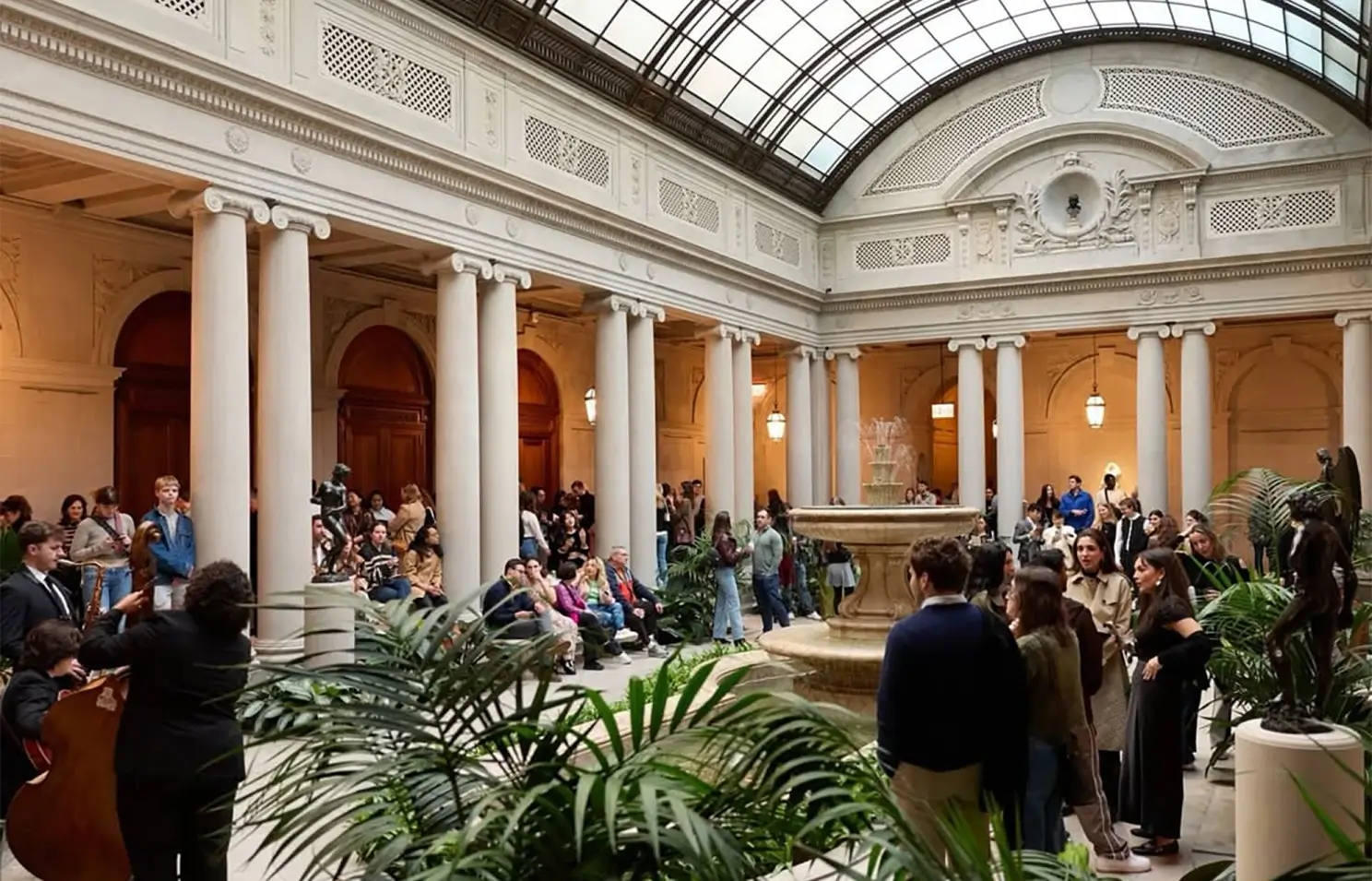 Visitors gather inside The Frick Collection’s restored Garden Court beneath a vaulted glass ceiling, surrounded by classical columns, indoor greenery, and a central fountain during a museum event