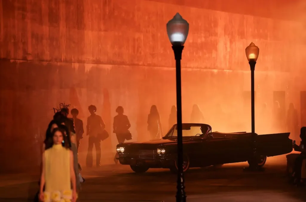 Atmospheric runway scene bathed in warm amber light featuring silhouetted figures, vintage street lamps, and a classic convertible creating a cinematic fashion presentation