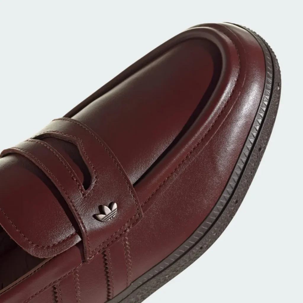 Close-up detail image of the adidas Originals HANDBALL SPEZIAL LOAFER PREMIUM in rich Fox Brown leather, emphasizing the smooth rounded moc-toe construction, penny-loafer inspired strap with metallic Trefoil accent, tonal stitching, and textured sidewall edge that gives the terrace-inspired silhouette a refined, elevated finish