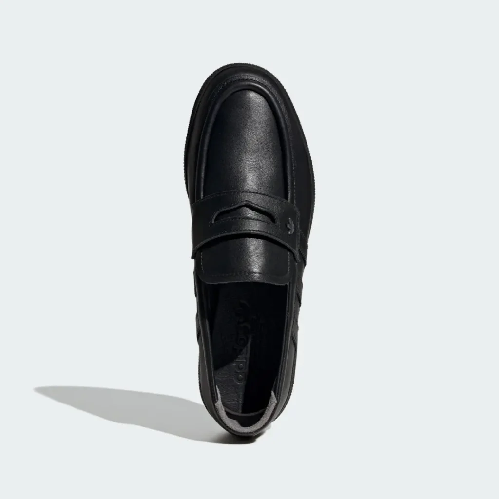 Top-down product image of the adidas Originals HANDBALL SPEZIAL LOAFER PREMIUM in black leather, showcasing its penny-loafer inspired strap, smooth rounded moc-toe construction, subtle Trefoil hardware detail, tonal Three Stripes along the sidewalls, and sleek monochromatic finish on a light grey background
