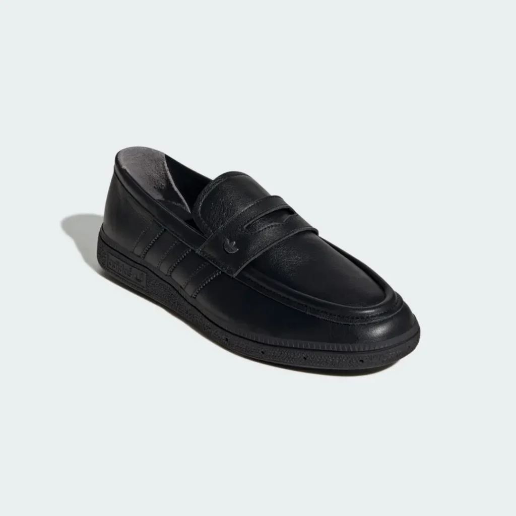 Three-quarter angled product image of the adidas Originals HANDBALL SPEZIAL LOAFER PREMIUM in black, highlighting its smooth leather construction, penny-loafer inspired strap with subtle Trefoil accent, tonal Three Stripes, rounded moc-toe detailing, and low-profile textured sole set against a minimal light grey backdrop