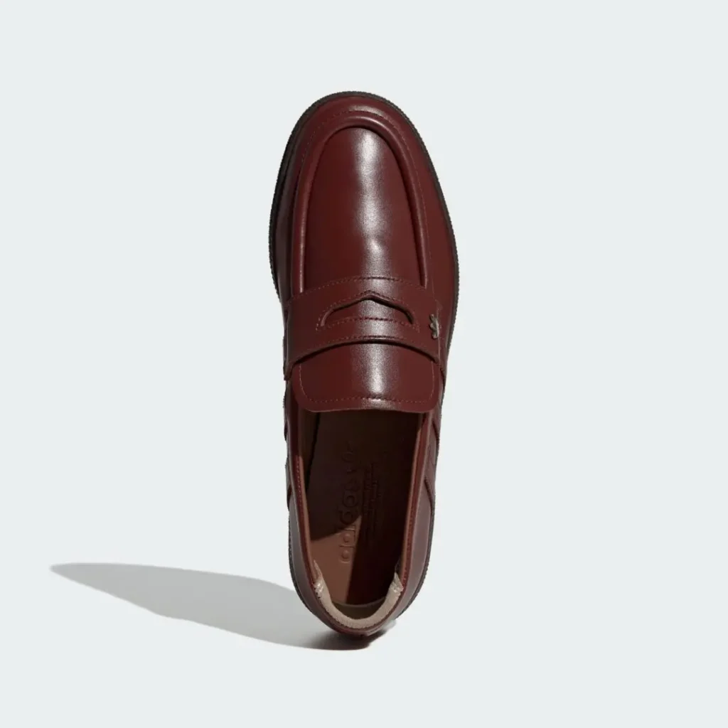 Top-down product image of the adidas Originals HANDBALL SPEZIAL LOAFER PREMIUM in rich Fox Brown leather, highlighting its penny-loafer inspired strap, smooth rounded moc-toe construction, subtle Trefoil hardware accent, tonal side striping, and sleek monochromatic finish set against a clean light grey background