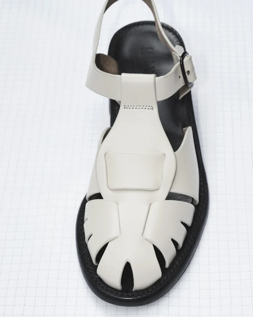 Minimal ivory leather fisherman sandal with sculpted cutout detailing, adjustable ankle strap, and black molded sole, photographed against a faint grid-pattern studio backdrop
