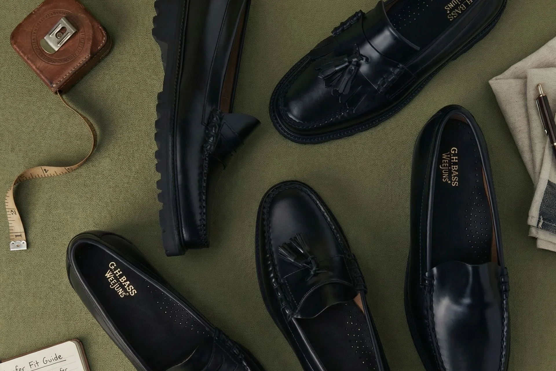 Flat lay of black G.H. Bass Weejuns loafers arranged across an olive fabric surface, featuring penny loafers, tassel loafers, and lug-sole variations alongside tailoring tools and accessories that reinforce the brand’s heritage menswear aesthetic