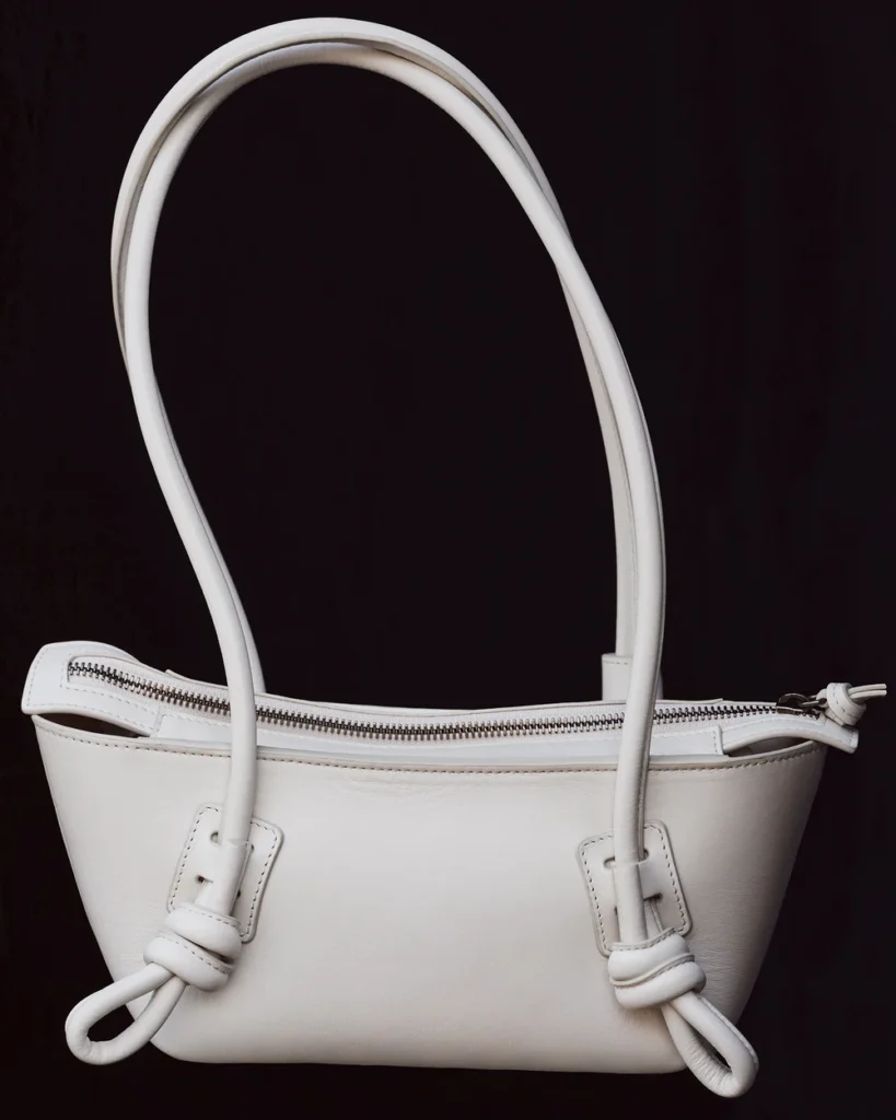 Structured white leather shoulder bag with elongated tubular handles, exposed silver zip closure, and sculptural knotted strap detailing, photographed against a deep black studio backdrop