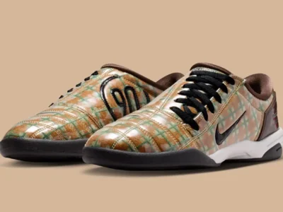 A pair of low-top Nike shoe is displayed against a neutral beige background, featuring a glossy plaid-patterned upper in muted gold, tan, and sage green tones with black laces and a black Swoosh. The quilted texture, retro indoor-soccer-inspired silhouette, and earthy palette give the shoes a distinctive lifestyle-meets-performance aesthetic with vintage appeal