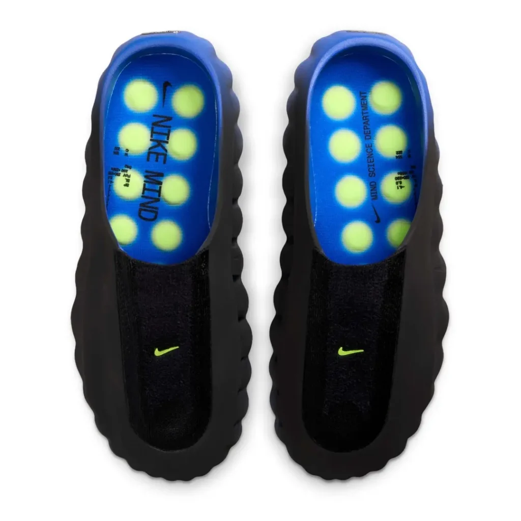 Top-down product image of the Nike Mind 001 Flyknit QS “Hyper Royal/Volt,” displaying both shoes side by side to highlight the interior construction and sensory footbed design. The vivid Hyper Royal insole features glowing Volt circular pressure zones beneath printed “NIKE MIND” and “MIND SCIENCE DEPARTMENT” branding, reinforcing the model’s neuroscience-inspired concept. A black Flyknit forefoot panel with a small Volt Swoosh contrasts against the sculpted dark foam shell, creating a futuristic mule silhouette focused on sensory engagement and pre-performance comfort
