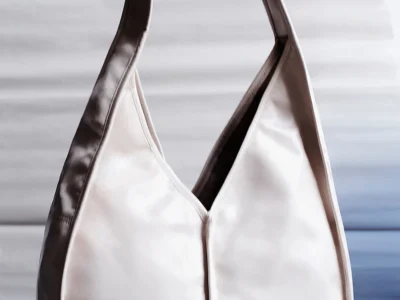 Minimal crescent-style shoulder bag in soft cream leather with deep espresso trim, sculptural curved strap, and folded center seam construction against a muted studio backdrop