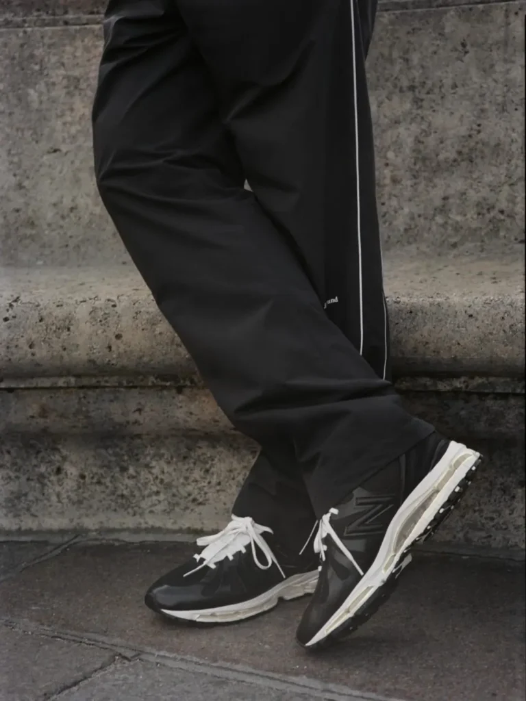 Lifestyle image of a person wearing black and cream New Balance 1890 by JJJJound sneakers with oversized white laces and visible ABZORB cushioning, styled with black technical pants against a dark stone urban backdrop
