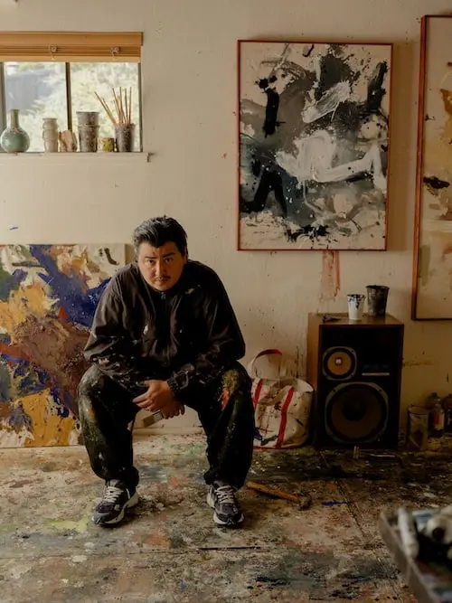 Lifestyle campaign image featuring an artist seated inside a paint-splattered studio while wearing the New Balance x BEAMS 2010 “Navy” shoe, surrounded by abstract canvases, speakers, and layered creative textures in a warm, lived-in workspace