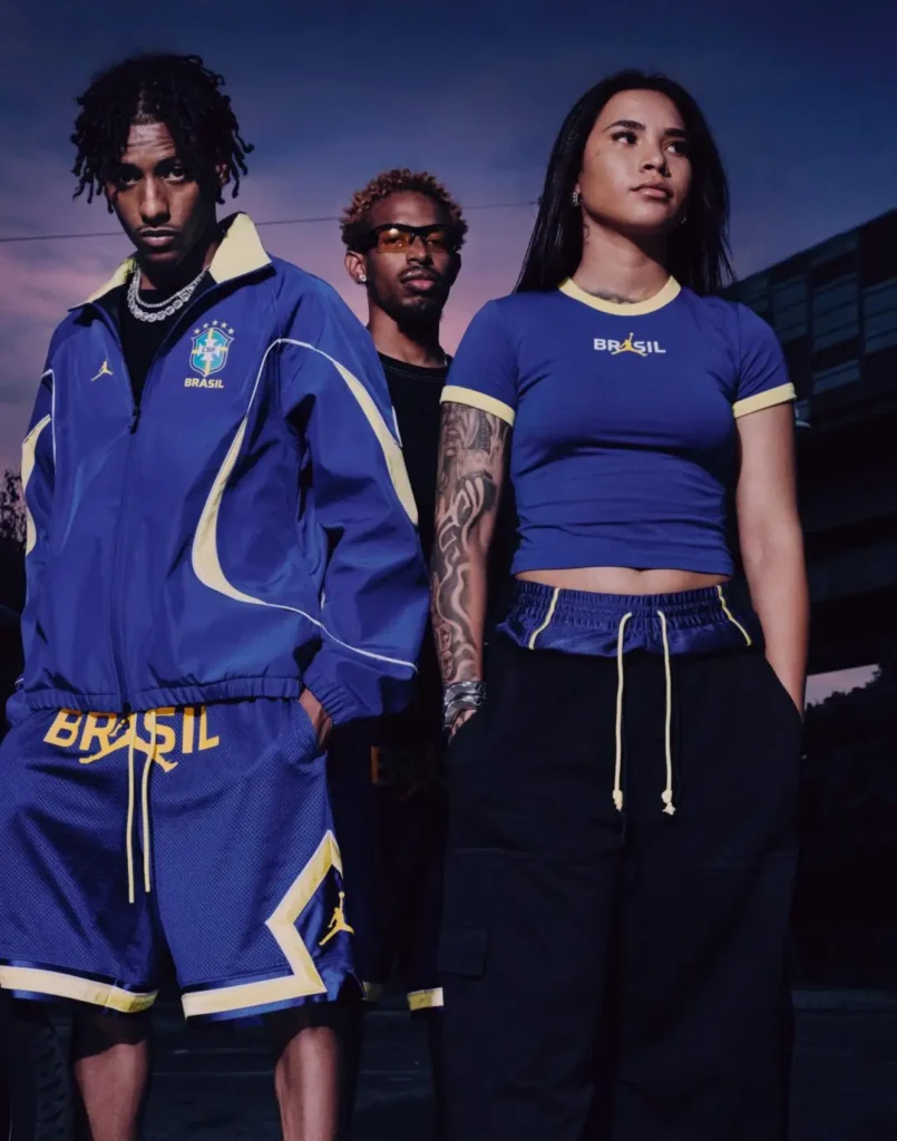 Nighttime campaign portrait featuring three models styled in Brazil-inspired Jordan Brand apparel against a dusky urban backdrop. The foreground includes a male model wearing a blue Brazil football jacket and matching mesh shorts with yellow accents and Jumpman branding, alongside a female model in a fitted blue “BRASIL” crop top paired with relaxed black drawstring pants. A third model stands between them in tinted glasses and layered streetwear accessories, reinforcing the collection’s fusion of football heritage, contemporary street fashion, and global sports culture