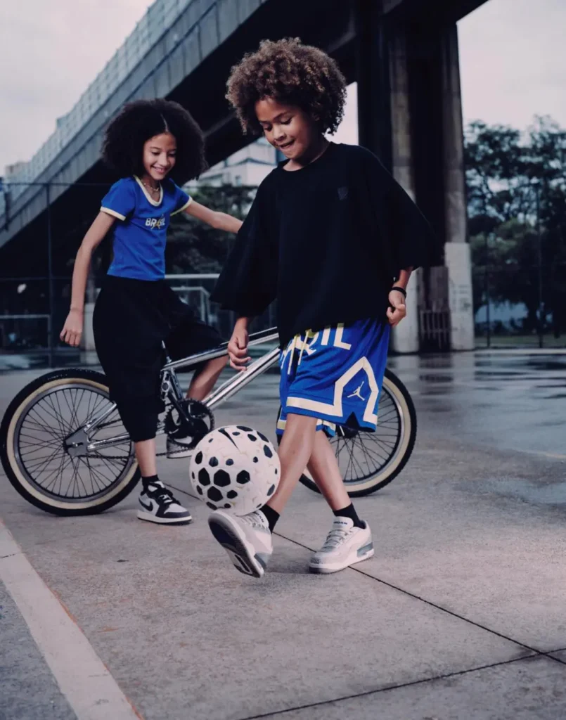 Two children styled in Brazil-inspired Jordan Brand apparel play casually with a football beside a bicycle in a modern urban setting at dusk. One child balances a black-and-white football while wearing oversized blue Brazil mesh shorts with yellow Jumpman detailing, a loose black top, and sneakers, while the other stands nearby in a fitted blue “BRASIL” T-shirt and cropped black pants. The cinematic scene blends youthful street culture, football energy, and contemporary sportswear styling against a moody city backdrop