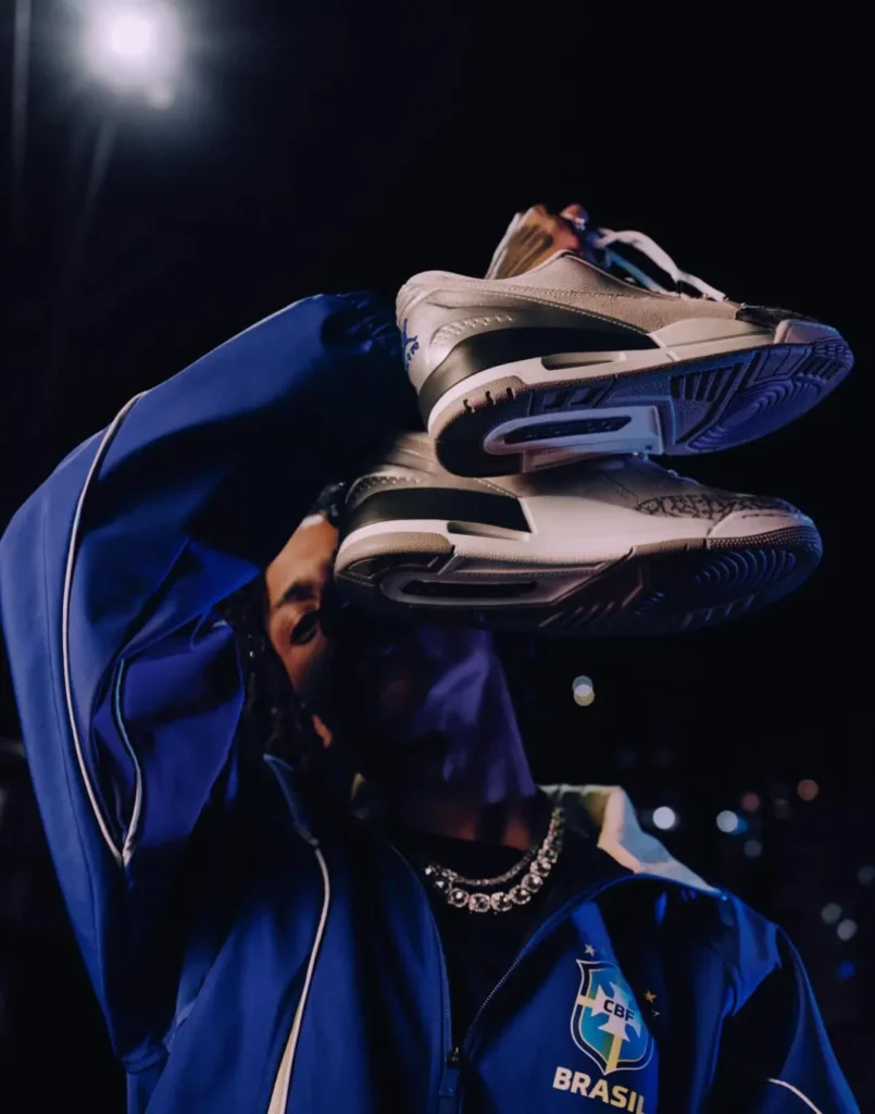 Nighttime campaign image of a model in a royal blue Brazil-inspired Jordan Brand track jacket holding a pair of white-and-black Air Jordan sneakers across their face under dramatic street lighting. The jacket features the Brazil football crest and “BRASIL” branding on the chest, while layered silver jewelry and blurred city lights reinforce the cinematic fusion of football culture, sneaker heritage, and contemporary streetwear styling